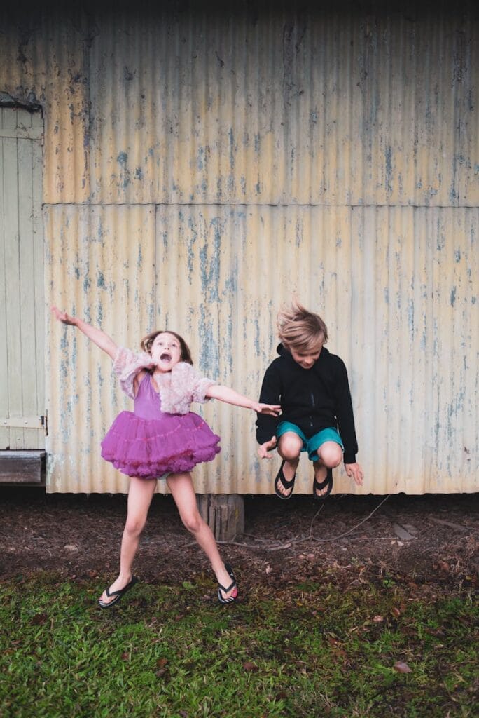 Two kids joyfully jump in front of a rustic wall, embodying pure excitement and fun.