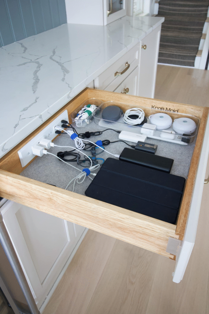 Illustration of a tidy kitchen drawer charging station with smartphones and a tablet. Charging cables run through grommets and plug into a realistic wall-mounted surge protector with USB ports above the drawer.